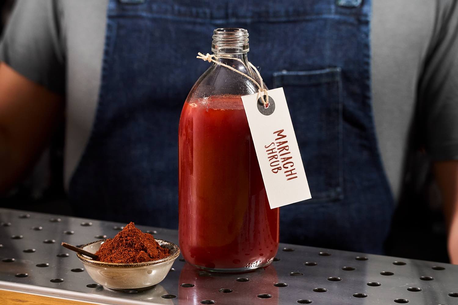 Una bottiglia di Mariachi Shrub con una ciotolina di chipotle. Mariachi Shrub è un premix a base di fragole, chipotle, miele e aceto di mele.