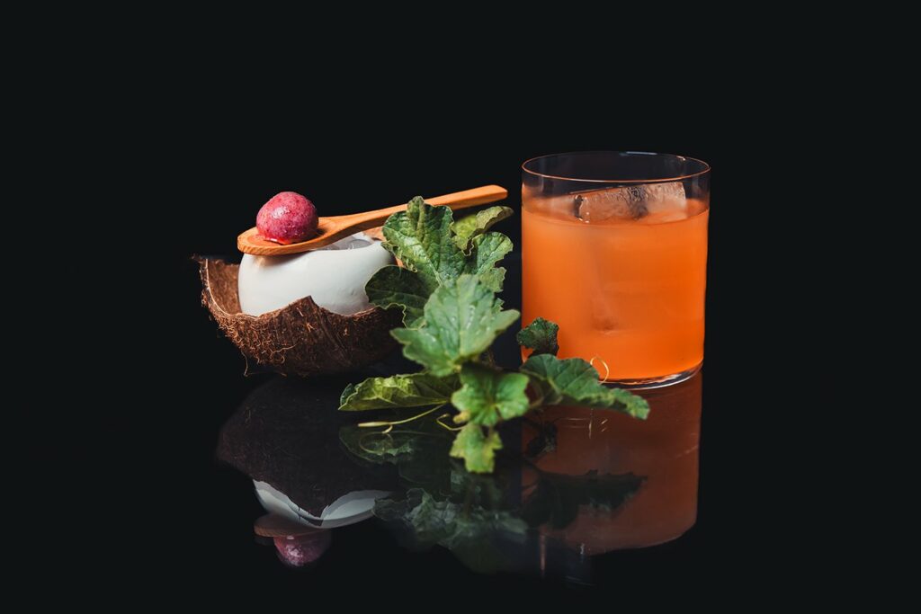 Un bicchiere di Halo Halo Cocktail con una cocottina in ceramica piena di ghiaccio su un pezzo di guscio di cocco. Sopra la cocottina un cucchiaino di legno con sopra una pallina di gelato al lampone.