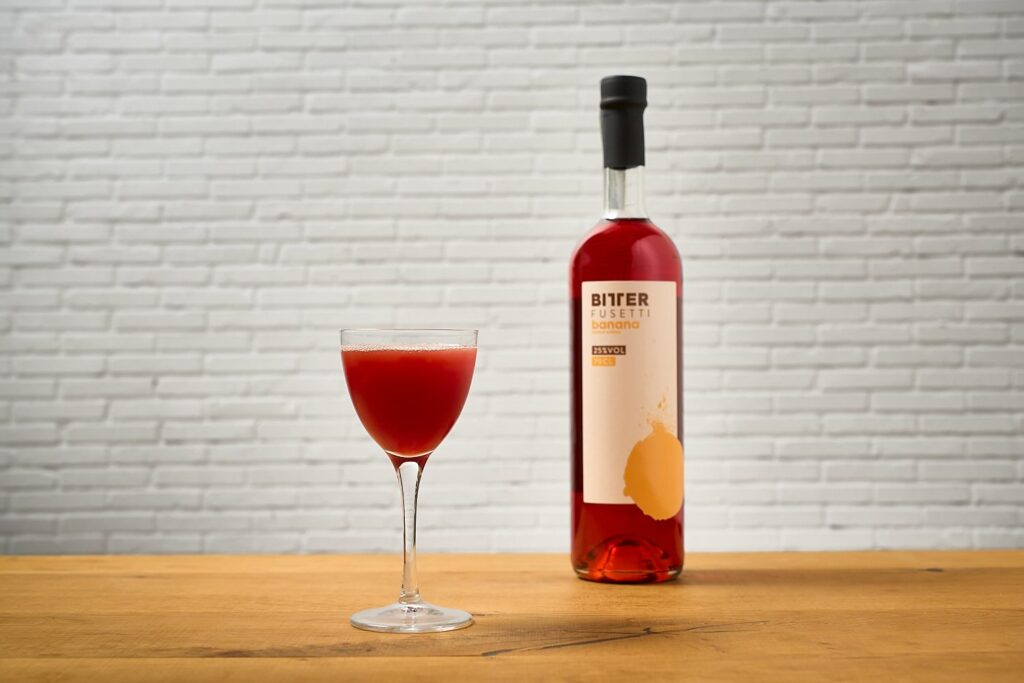 il cocktail Fusettino in una coppetta su un tavolo di legno. Sullo sfondo una bottiglia di Bitter Fusetti alla Banana