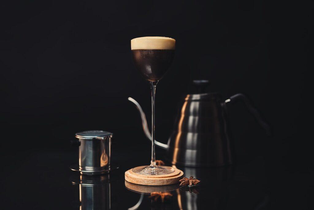 Il cocktail Vietnamese Moretta in una coppetta da cocktail classica dallo stelo alto. sullo sfondo un tipico bollitore con collo a cigno e il Phin per preparare il caffè vietnamita.
