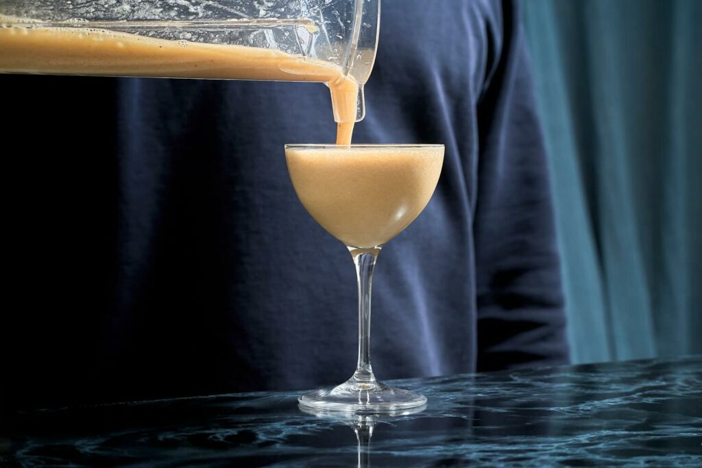 su un banco bar in marmo scuro, una coppetta contenente il cocktail Fermented Banana Daiquiri versato dalla campana di un frullatore.