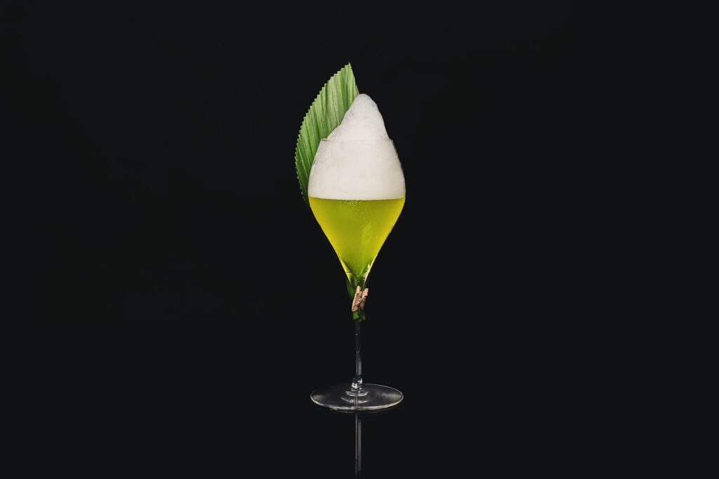 Il cocktail French' doutre-mer su uno sfondo nero. Il cocktail è servito in un calice a tulipano con una foam in superficie ed è decorato con una foglia di banano.