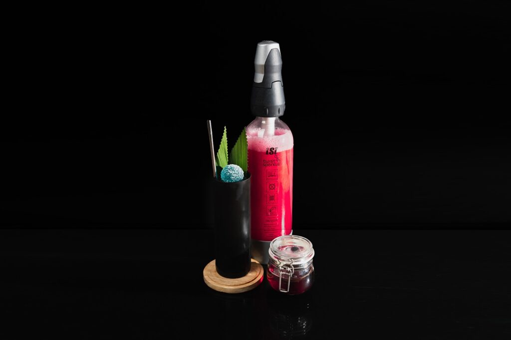 In un bicchiere nero il twist del cocktail doctor funk decorato con una foglia di banano tagliata e con la caramella gommosa all’anice varnelli. Dietro al cocktail un sifone “Twist’n Sparkle” con all’interno la soda alla granatina home made.