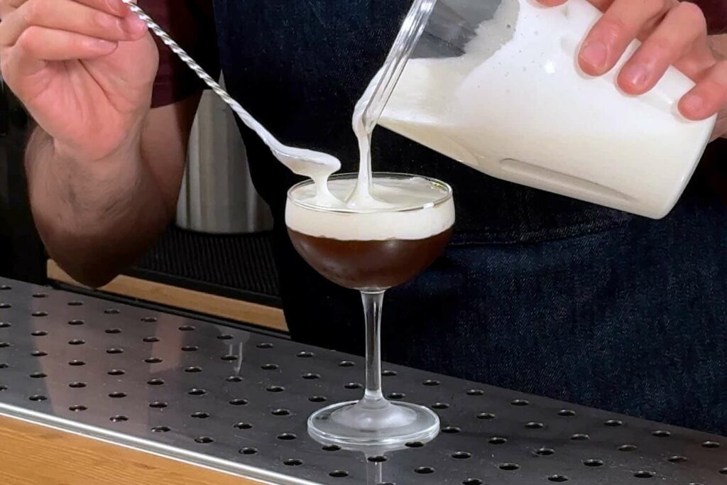 Su un banco bar una coppetta con un cocktail al caffè sul quale viene versata una foam al kefir direttamente dal contenitore di un milkshake mixer