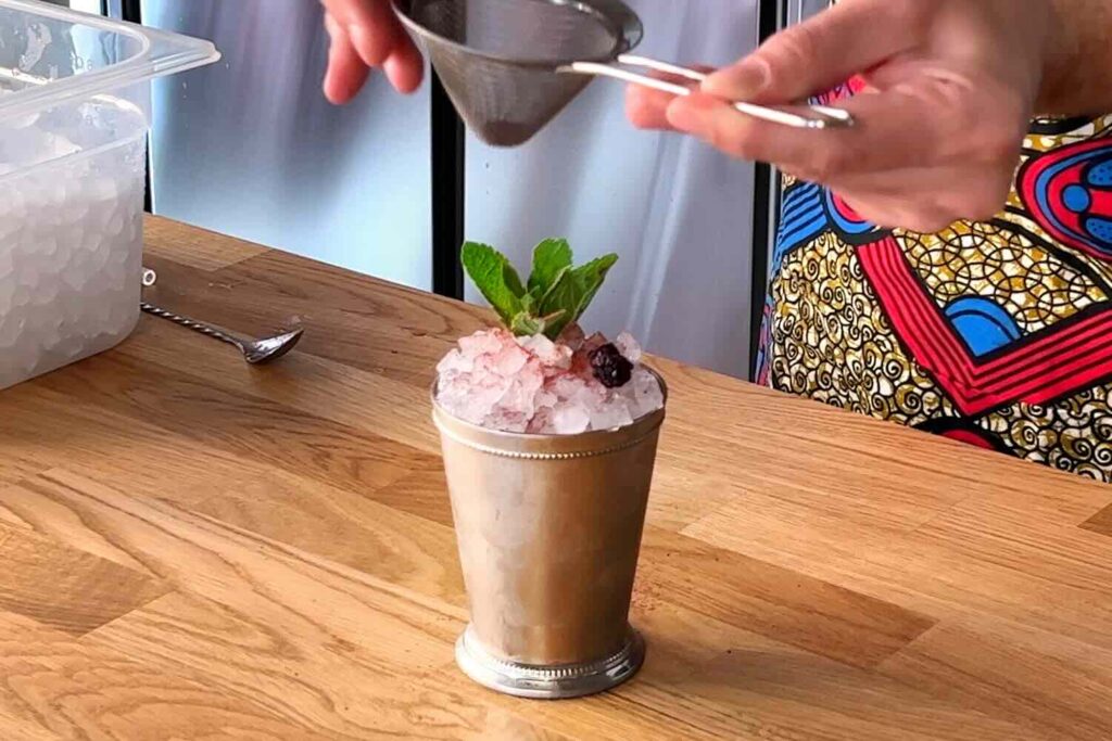 Su un banco bar in legno il cocktail "meglio le more". Il cocktail si presenta nella classica julep mug in acciaio con un cumulo di ghiaccio tritato che fuoriesce. Il cocktail è decorato con un ciuffetto di menta, una mora essiccata e polvere di more essiccate