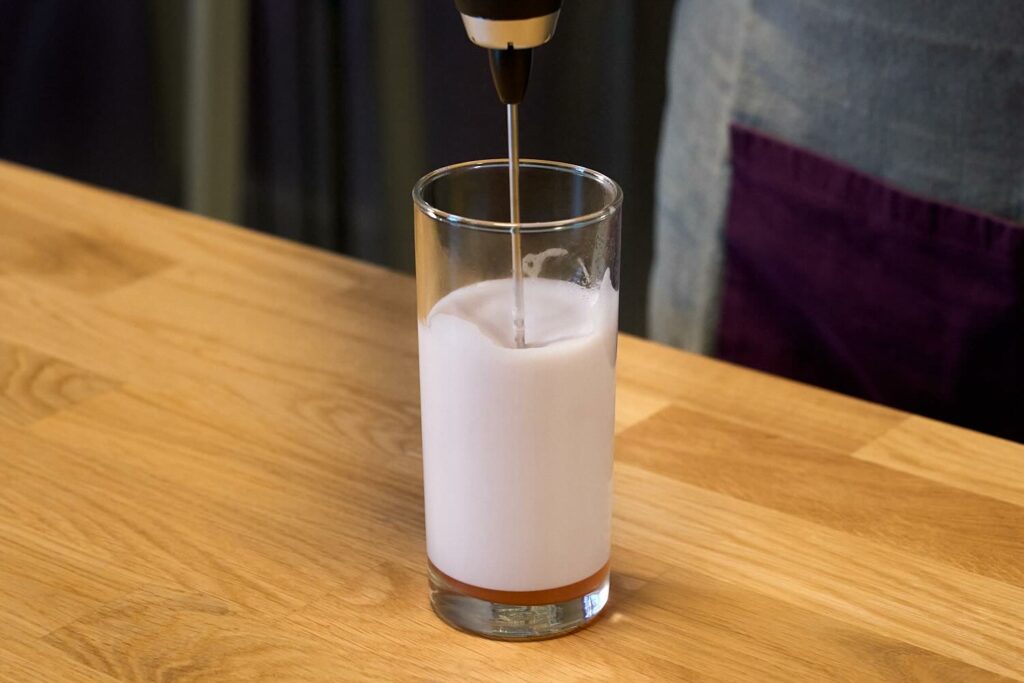 Su un banco bar un bicchiere alto di vetro con all'interno il velluto di fragole. Nel liquido è immerso il frullino monta latte che sta areando la schiuma