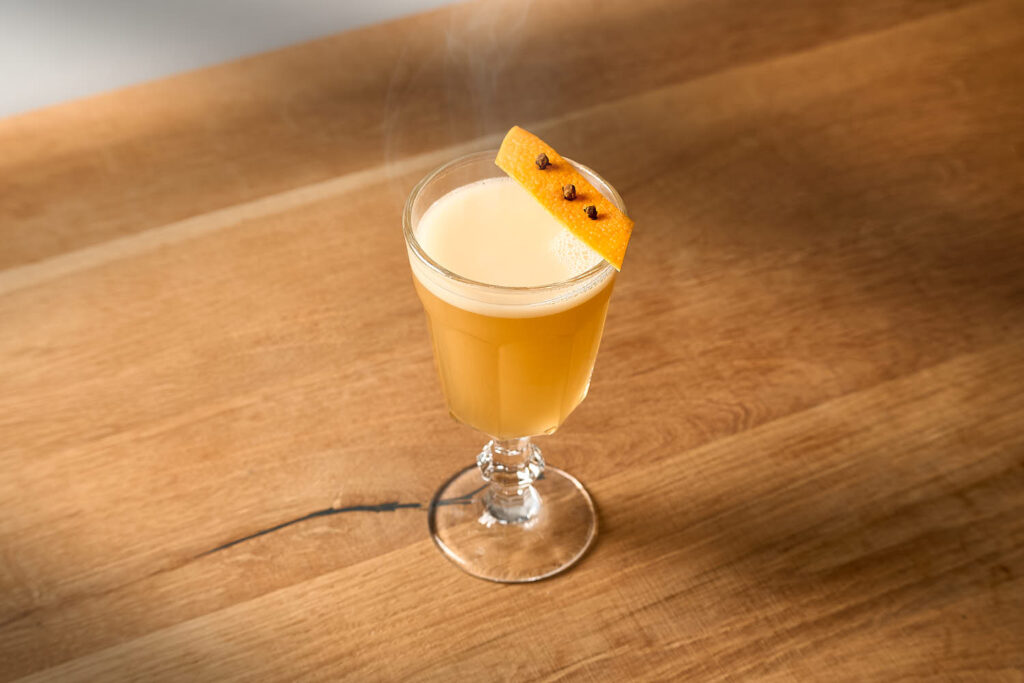 Su un tavolo in legno il cocktail caldo Hot Tiger Milk. Il cocktail è servito in una coppetta alta in stile vintage ed è decorato con una scorza di arancia e 3 chiodi di garofano conficcati.