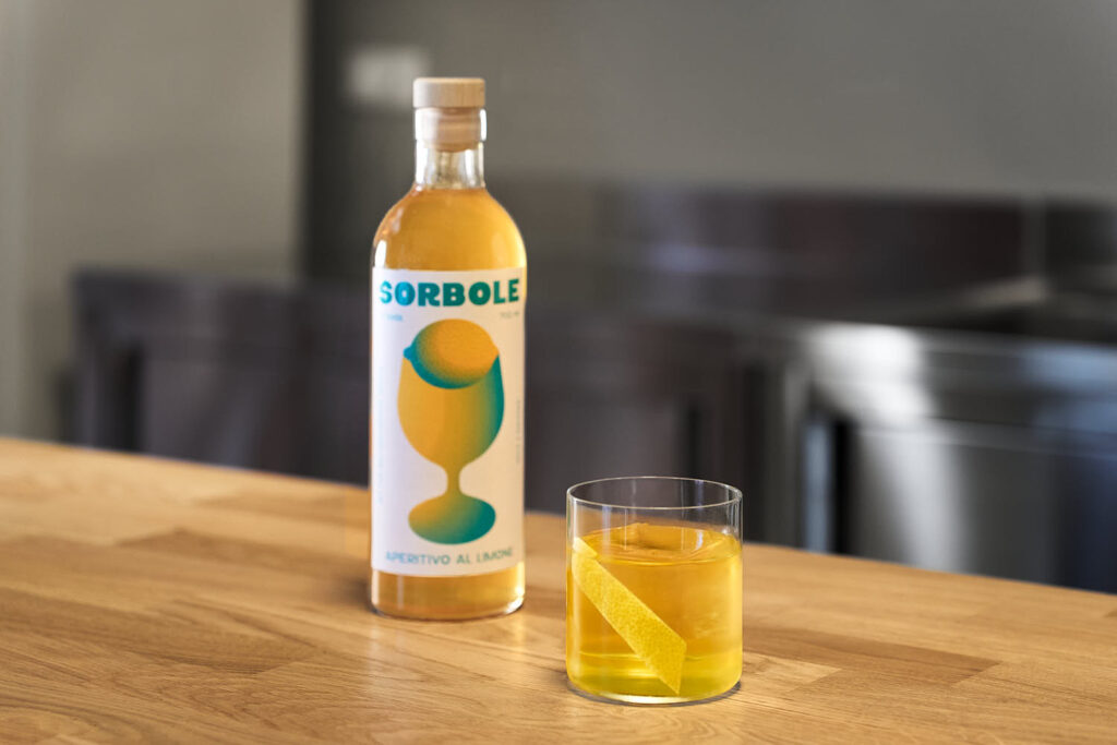 Su un banco bar Il cocktail Negroni Giallo servito in un bicchiere tipo tumbler basso con un chunk di ghiaccio e decorato con una scorza di limone. Accanto al cocktail una bottiglia di Sorbole, aperitivo al limone.