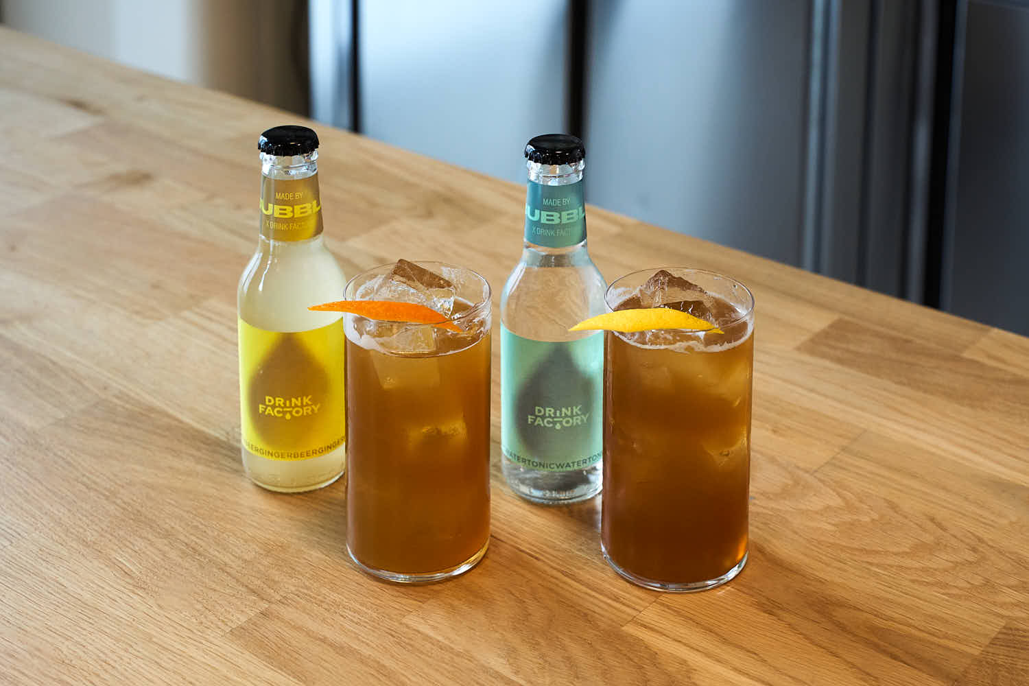 Su un banco bar di legno, due bicchieri da Long drink contenenti due cocktail analcolici, preparati con il pre mix Indian Chai coffee Cordiali. Accanto ai due cocktail, una bottiglietta di Ginger Beer e una di tonica prodotte da Bubble Bespoke.