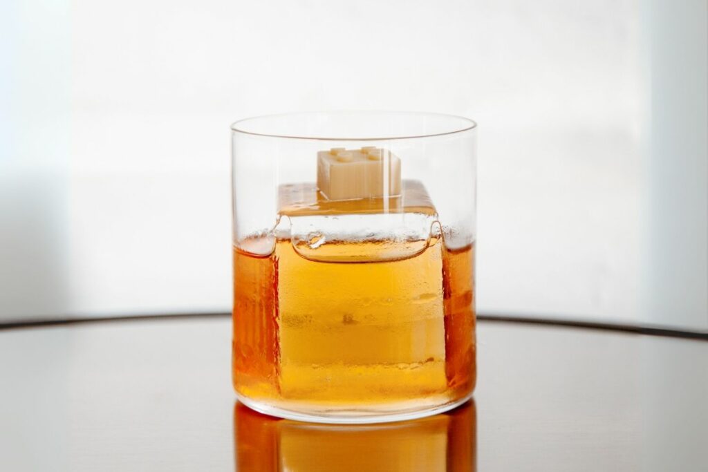 Su un tavolo a specchio il cocktail Monkey Glam. Il drink è di colore dorato ed è servito su un chunk di ghiaccio in un tumbler basso dall'aspetto minimale. La decorazione consiste in un mattoncino lego realizzato con cioccolato bianco.