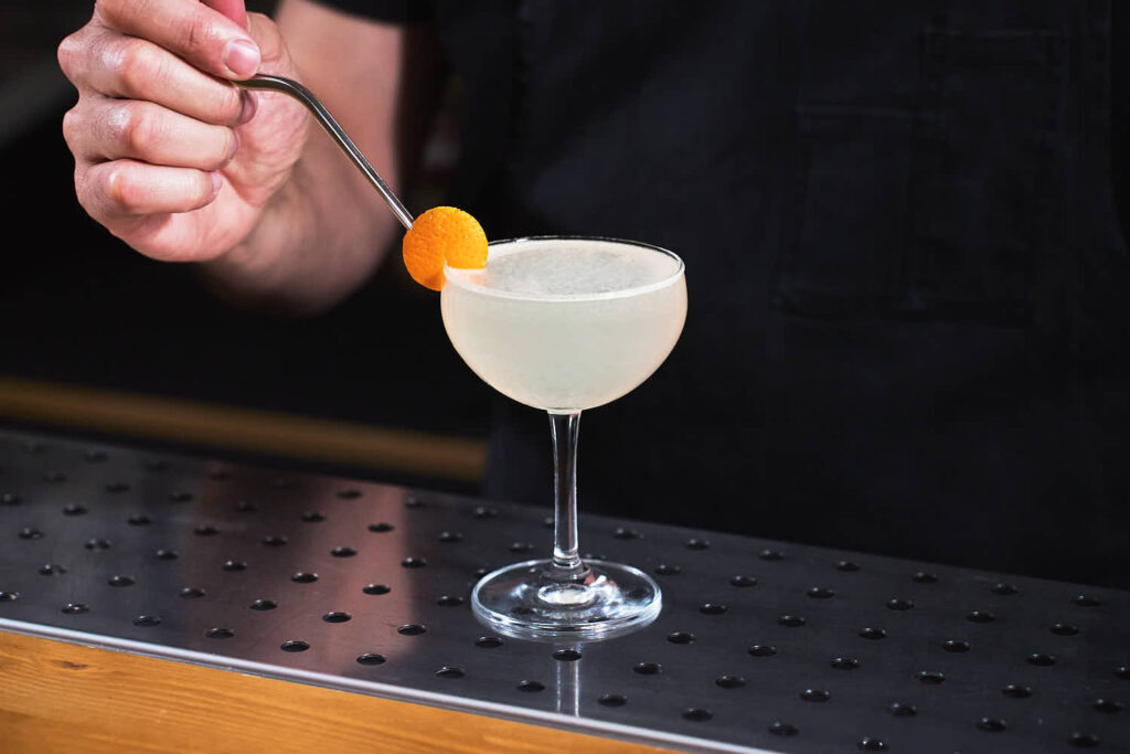 Su un banco bar una coppetta da cocktail contenente il cocktail Corpse Reviver n°2 preparato con Gin, americano cocchi, succo fresco di limone, triple sec e assenzio. Il cocktail è decorato con una scorza di arancia tagliata tonda e incastrata sul bordo del bicchiere.