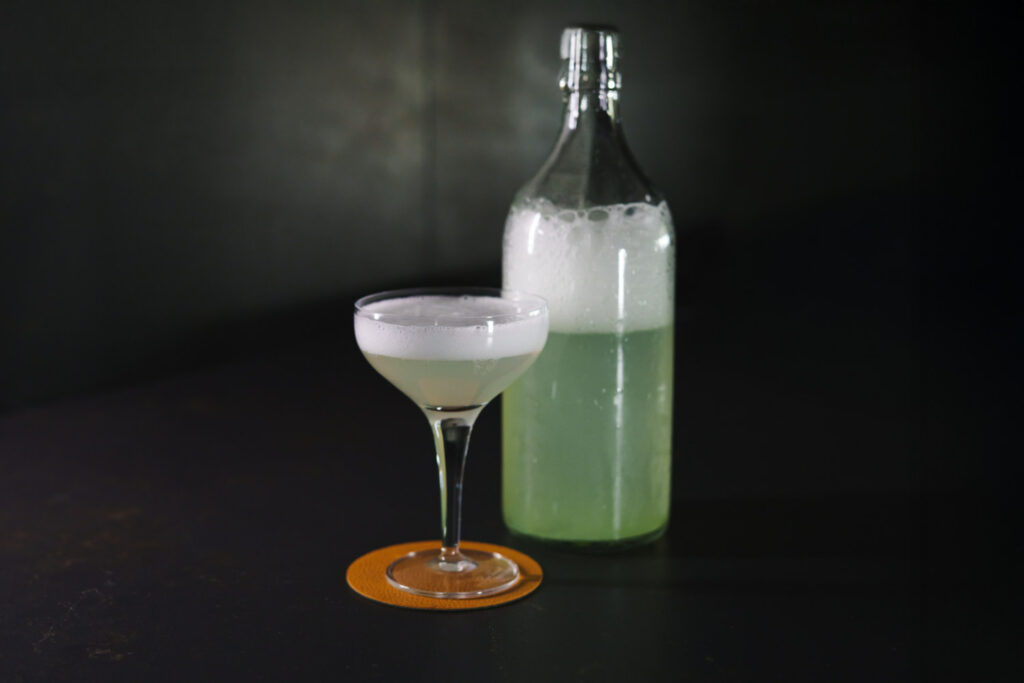 su un tavolo una bottiglia e un bicchiere da cocktail contenenti un fermentato di fiori di sambuco utilizzato come ingrediente nel cocktail "hugo dal futuro"