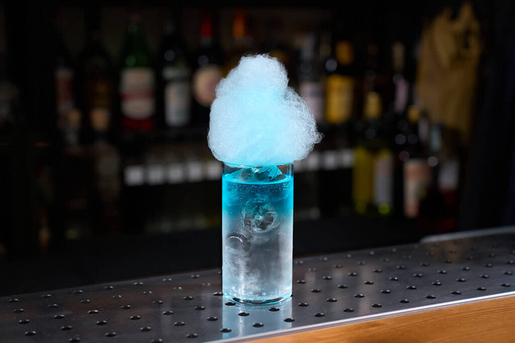 Su un banco bar il cocktail Mistico Blu. Il cocktail, servito in un bicchiere da long drink con ghiaccio) è trasparente nella parte bassa e leggermente colorato di blu nella parte superiore. La decorazione è una nuvola di zucchero filato azzurro