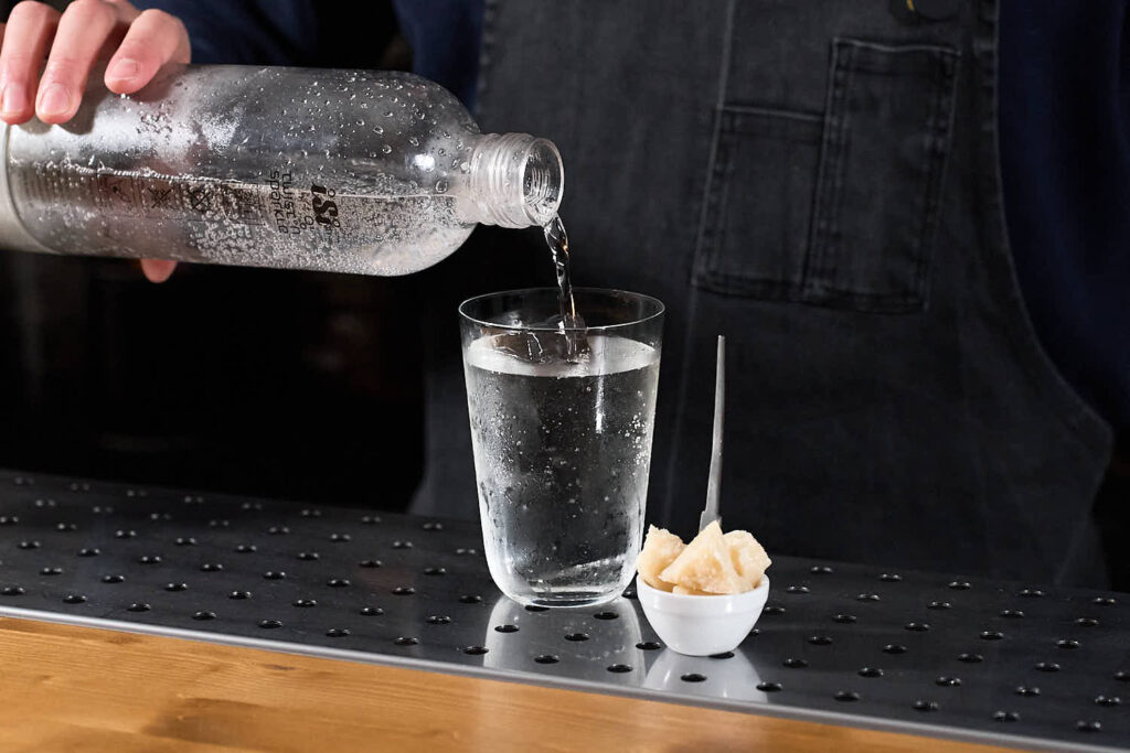 Su un banco bar un cocktail con soda alla pera servito in un bicchiere da long drink. Accanto al cocktail una piccola ciotola in ceramica bianca contenente dei pezzetti di Parmigiano reggiano. Si intravede la mano del barman che versa la soda nel bicchiere da un sifone "Twist'n Sparkle"